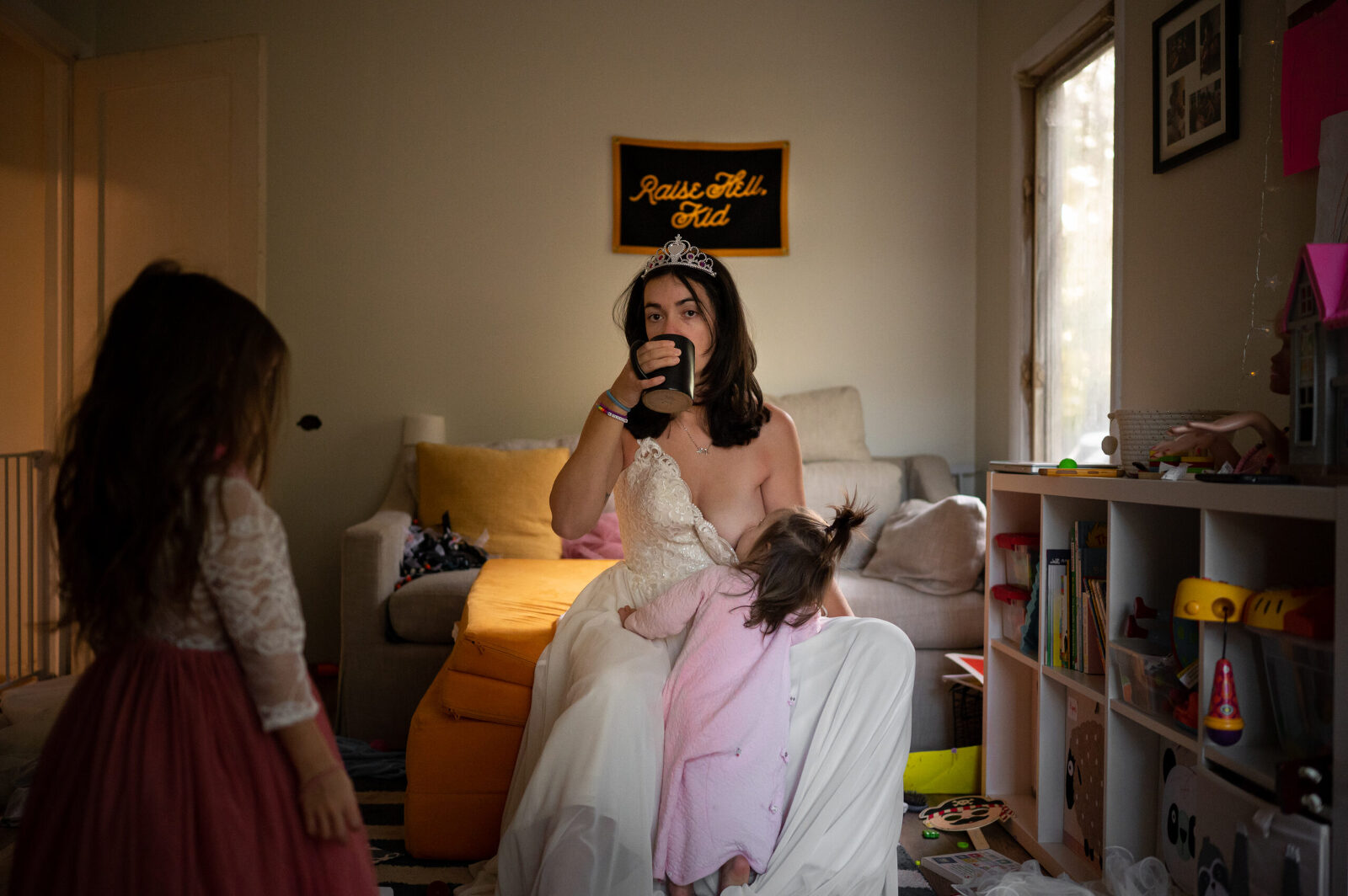 A mother wearing her wedding dress and kids tiara sitting on nugget drinking coffee while toddler breastfeeds